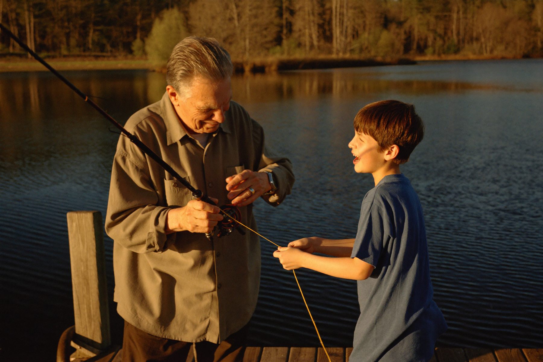 A grandfather showing his grandson how to tie a fishing line at the end of a dock at golden hour.