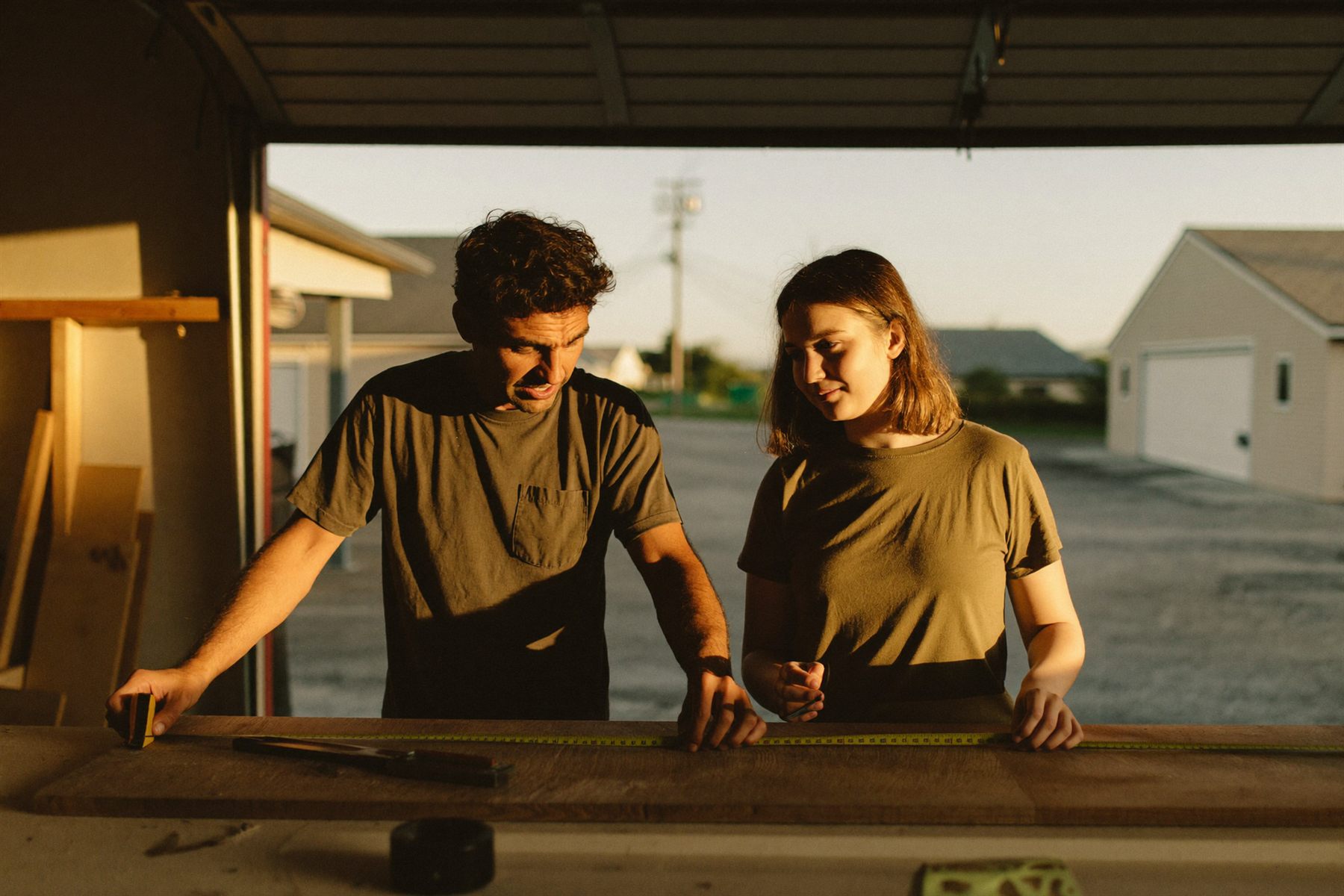A father and his teenage daughter measuring a board together at a workbench in a garage.