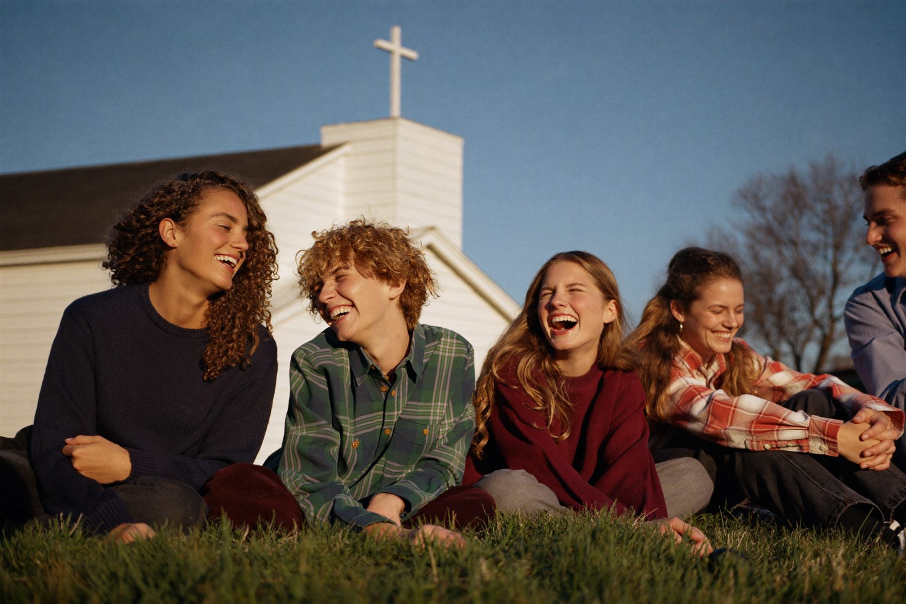 A youth leader and four high school students laughing together on the grass beside a white country church.