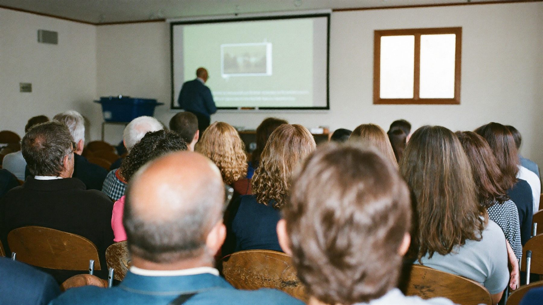 A speaker addressing a seated group of adults in a church fellowship hall, seen from the back of the room.